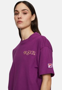 Paarse katoenen t-shirt met goud geborduurde "R.S.V.P." tekst en een logo patch op de mouw. Losse pasvorm met korte mouwen.