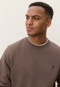 Sweat-shirt marron avec col et poignets côtelés, arborant un petit logo noir "II" sur le côté gauche. Texture lisse, coupe décontractée.