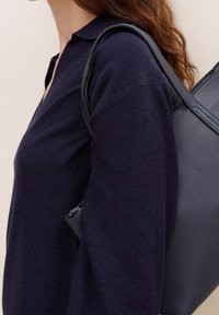 Pullover blu navy con colletto indossato sopra una borsa a tracolla texturizzata. Il tessuto appare morbido e lavorato a maglia con una vestibilità comoda.