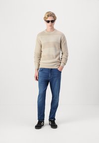 Maglione beige a maglia con righe orizzontali, jeans blu e sneakers nere. Modello con occhiali da sole neri in piedi su uno sfondo bianco.