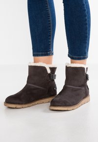 Ruskih nubuk čizama do gležnja s lažnom krznom oblogom, detaljem sa remenom i dugmetom, te teksturiranim gumiranim đonom. U kombinaciji s tamnim trapericama.