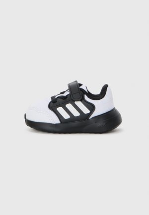 Baskets noir et blanc avec une tige en maille, une sangle Velcro et une semelle noire. Présentent trois rayures blanches sur le côté.