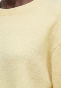 Gros plan d'une personne portant un pull tricoté doux jaune pâle avec un col rond et des cheveux courts visibles.