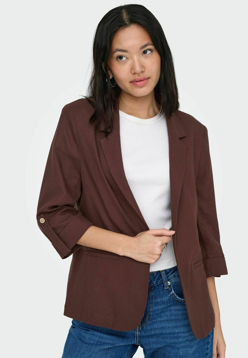Jeune femme aux cheveux noirs raides portant un blazer marron par-dessus un haut blanc et un jean bleu, posant devant un fond blanc uni.