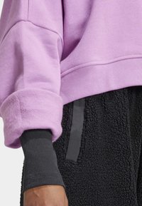Fialová mikina so nadýchaným manžetom a textúrovanou čiernou chlpato látkou v páse, s hladkým čiernym akcentom pozdĺž strany.
