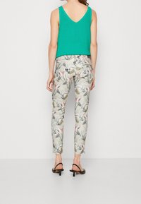 Femme portant un haut sans manches vert, un pantalon beige avec des motifs d'oiseaux et de feuilles, et des sandales à talons noires, se tenant contre un fond blanc uni.