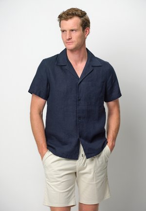 Dunkelblaues, kurzärmeliges Leinenhemd mit Camp-Kragen, einer Brusttasche und vorderer Knopföffnung, kombiniert mit hellbeigen Shorts.