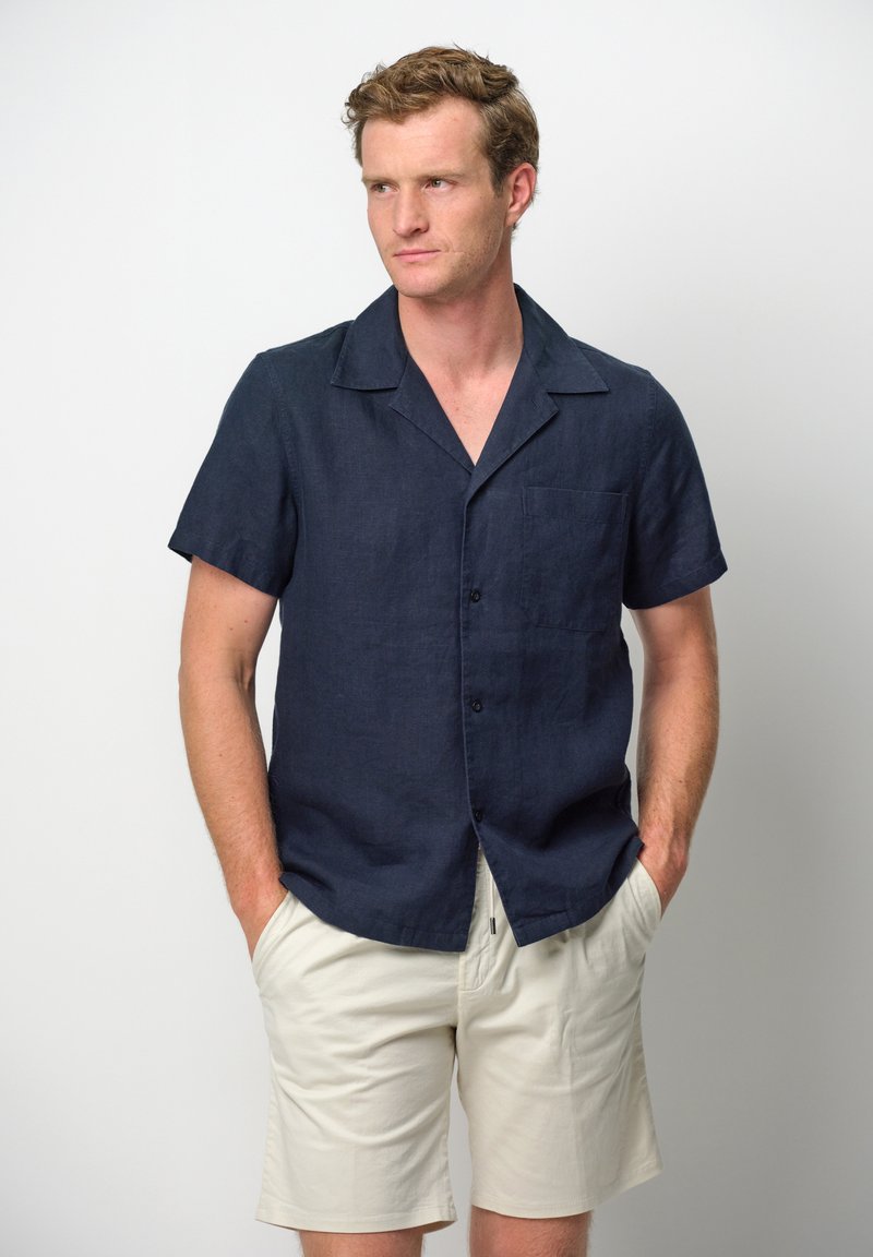 Camicia in lino a maniche corte di un blu navy scuro con colletto da campeggio, taschino sul petto e chiusura frontale con bottoni, abbinata a pantaloni corti beige chiaro.