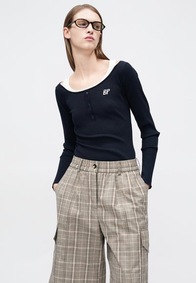 Haut à manches longues côtelé marine avec encolure arrondie crème et boutons, associé à un pantalon à jambes larges beige à carreaux avec poches latérales.