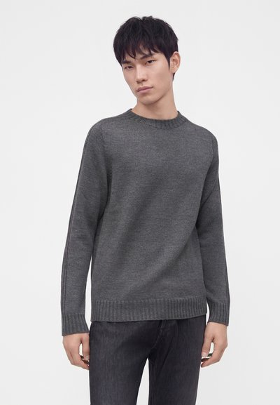Jeune homme aux cheveux courts noirs portant un pull en maille gris foncé uni et un pantalon noir sur fond blanc uni.