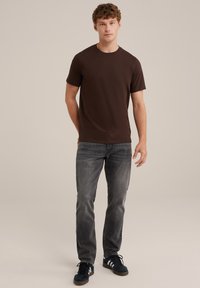 Camiseta marrón de manga corta en un corte clásico, combinada con pantalones vaqueros oscuros. El conjunto se completa con zapatillas azul marino con rayas blancas.