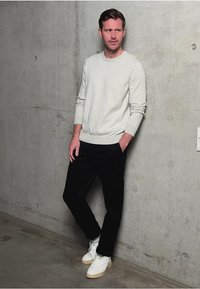 Helles graues, strukturiertes Sweatshirt, schwarze Hose, weiße Sneaker mit grünen Akzenten. Person steht gegen eine Betonwand.