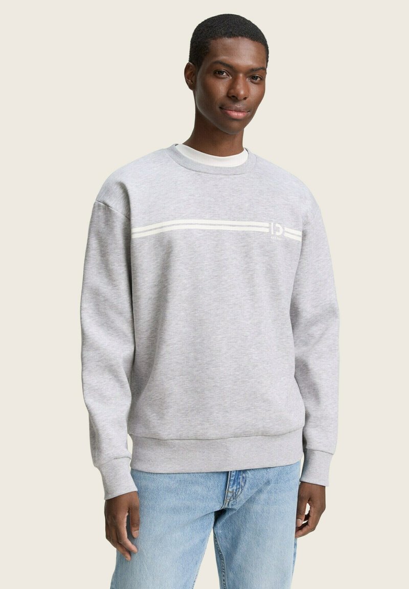 Pilkas džemperis, pagamintas iš medvilnės ir sintetinių pluoštų, su apvalia iškirpte, ilgomis rankovėmis ir baltais horizontaliais dryžiais krūtinėje. Klasikinio kirpimo.