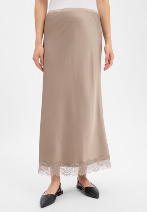 Femme portant une longue jupe beige avec une bordure en dentelle à l'ourlet et des ballerines slingback noires pointues, debout sur fond blanc.