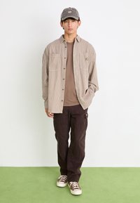 Striped beige corduroy shirt, brown t-shirt, brown cargo pants, and khaki cap with logo. White sneakers. Plain background with green floor.