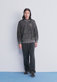 O persoană tânără cu păr lung și negru poartă un pulover cu model închis, pantaloni negri largi și adidași negri, stând cu spatele la un perete luminos, simplu.