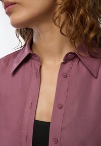 Camicia a maniche corte con colletto in tessuto mauve, caratterizzata da una chiusura frontale con bottoni e una texture liscia; indossata sopra un top nero.