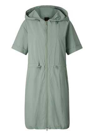 Vrouw met oversized zwarte zonnebril, muntgroene capuchonjurk met rits, bijpassende sneakers, houdt een zwarte rugzak naast zich.