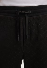 Zwarte joggingsbroeken met een gewatteerde textuur, voorzien van een zwarte elastische tailleband en trekkoord. Het patroon bevat reliëfdesigns.