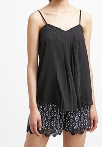 Svart plisserad linne med tunna axelband, parat med mönstrade svarta shorts med spetsdetalj i fållen. Slät tygtextur.