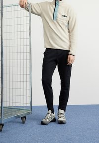 Pullover in pile beige con dettagli teal, collo a zip e tasca sul petto. Pantaloni cargo neri con tasche e scarpe sportive grigie. Griglia metallica nelle vicinanze.