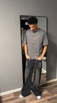 Mladý muž stojí s rukama v kapsách, má na sobě šedé triko Oakley, široké černé džíny a bílé boty před zrcadlem na zdi.