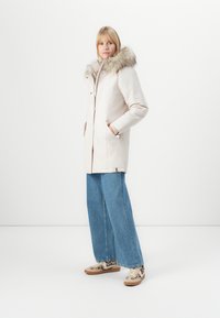 Parka de couleur claire avec garniture en fausse fourrure, dotée d'une fermeture éclair, de poches latérales et de manches longues, associée à un jean large et des chaussures à motifs.