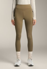 Olajzöld, magas derekú, testhez simuló leggings, rugalmas anyagból készült. Kiemelt varrásokkal és cipzáras zsebekkel rendelkezik. Fehér sportcipőt viselnek.