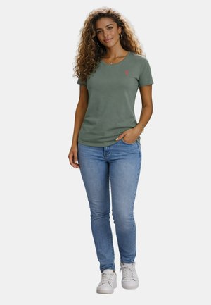 Woman with curly hair wearing a green short-sleeve t-shirt, light blue jeans, white sneakers, standing with one hand in pocket.