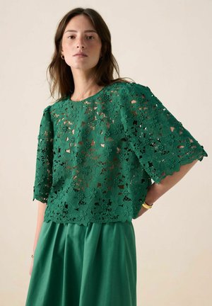 Green lace top featuring floral patterns, short sleeves, and a round neckline, paired with a matching green skirt. Background is neutral.