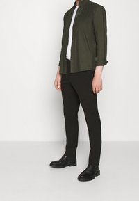 Camicia a maniche lunghe verde scuro indossata sopra una t-shirt bianca, abbinata a pantaloni neri slim-fit e stivaletti neri. Tessuto liscio, design con bottoni.