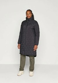 Sieviete, tērpta melnā quiltētā garajā mētelī, olīvzaļās biksēs un baltos sporta apavos, stāv pretī vienkāršai fonam.