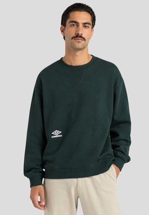 Uomo con capelli scuri e baffi che indossa una felpa Umbro verde scuro e pantaloni beige chiaro su sfondo grigio semplice.