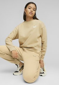 Felpa beige e pantaloni della tuta realizzati in tessuto morbido, con un piccolo logo bianco e cuciture a contrasto, abbinati a sneakers nere e bianche.