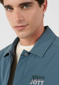 T�ürkisfarbene, leichte Jacke mit Kragen und gesticktem Logo auf der Brust, kombiniert mit einem cremefarbenen Strickpullover darunter.