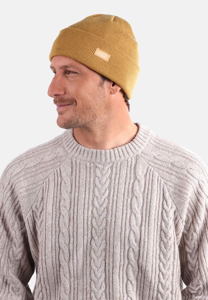 Bonnet en tricot ribé jaune avec une étiquette, porté par un homme dans un pull beige en tricot câble, arborant des motifs texturés et un design à couronne ronde.