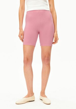 Vrouw draagt geribbelde roze biker shorts, crème platte schoenen en een witte mouwloze top, zichtbaar van midden van het bovenbeen tot aan de taille.