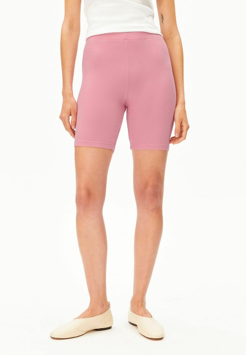 Donna che indossa pantaloncini da biker rosa a coste, scarpe piatte color crema e una canotta bianca senza maniche, mostrata dalla metà coscia alla vita.