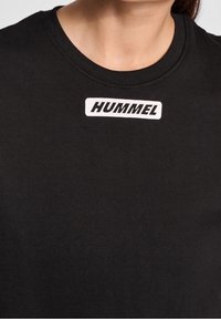 Sweat-shirt noir avec un col rond et des poignets côtelés. Présente un patch logo rectangulaire blanc avec "HUMMEL" en lettres noires en gras.
