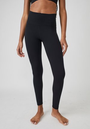 Mujer con leggings negros de cintura alta y largo completo, de pie descalza con las manos relajadas a los lados, sobre un fondo liso.