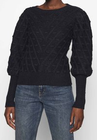 Svart stickad tröja med en strukturerad mönster med upphöjda bobbles, rund halsringning, och åtsittande ribbade manschetter och nederkant. Matchas med blå jeans.