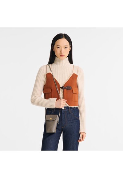 Brown suede cropped vest with front pockets, worn over a cream ribbed turtleneck, paired with dark blue jeans and a gray crossbody bag.