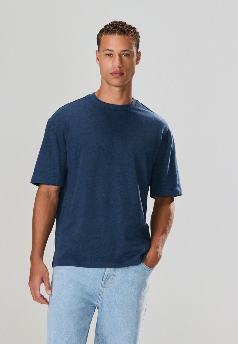 Jonge man met krullend haar, een marineblauw t-shirt en lichtblauwe jeans aan, staand met één hand in zijn zak tegen een effen achtergrond.