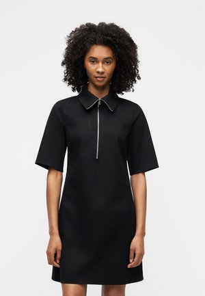 Mujer con cabello rizado que lleva un vestido negro de manga corta con cuello y cremallera frontal, de pie contra un fondo claro y liso.