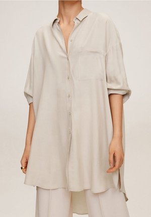 Chemise oversize beige clair à boutons, avec une poche poitrine, des manches courtes retroussées et une texture douce, avec des fentes latérales pour faciliter les mouvements.