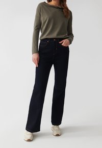 Femme portant un haut à manches longues vert olive, un jean droit taille haute bleu foncé et des baskets blanches, se tenant devant un fond uni.