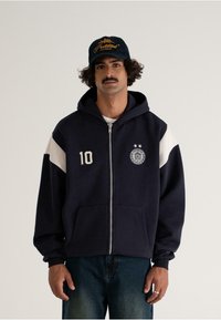 Man with curly hair and mustache wearing navy zip-up hoodie with "10" and emblem, white-striped sleeves, and a black cap with yellow text.