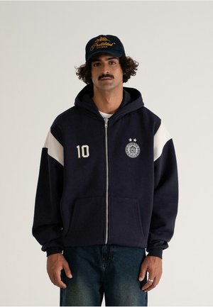 Man with curly hair and mustache wearing navy zip-up hoodie with "10" and emblem, white-striped sleeves, and a black cap with yellow text.