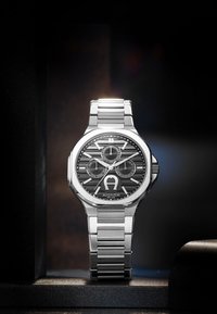 Montre en acier inoxydable argenté avec un cadran noir à motif, trois sous-cadrans et un logo bien en évidence. Elle dispose d'un bracelet à maillons et d'un boîtier aux angles marqués.