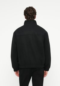 Μαύρο fleece μπουφάν με ψηλό λαιμό, ελαστικούς καρπούς και χαλαρή εφαρμογή. Ο ύφασμα είναι λείος χωρίς ορατά σχέδια ή διακοσμήσεις.